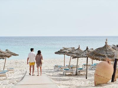 Ein Paar geht am Strand auf einem Holzsteg zwischen Strohmatschirmen Richtung Meer.