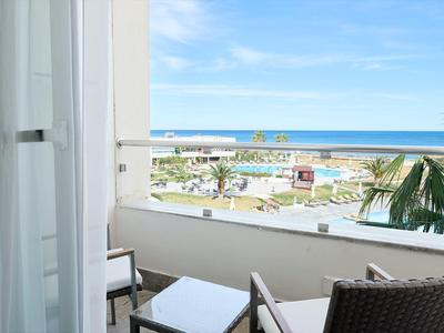 Balkon mit Stühlen und Tisch, Blick auf Palmen, Pool und Meer unter blauem Himmel.