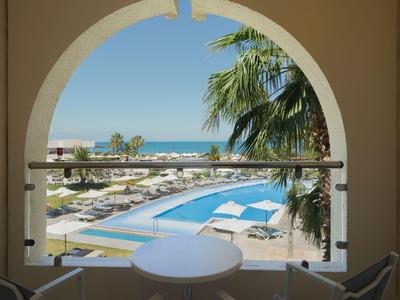 Blick durch einen Bogen auf Pool, Sonnenschirme, Palmen und Meer bei klarem Himmel.