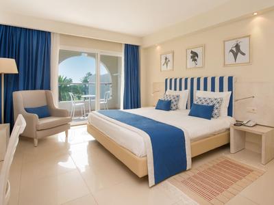 Camera d'hotel luminosa con letto matrimoniale, tende blu e balcone con vista mare.