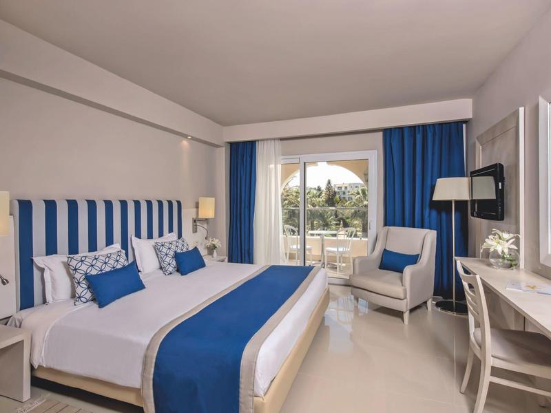 Camera d'hotel con letto matrimoniale, tende blu e balcone con vista sul giardino.