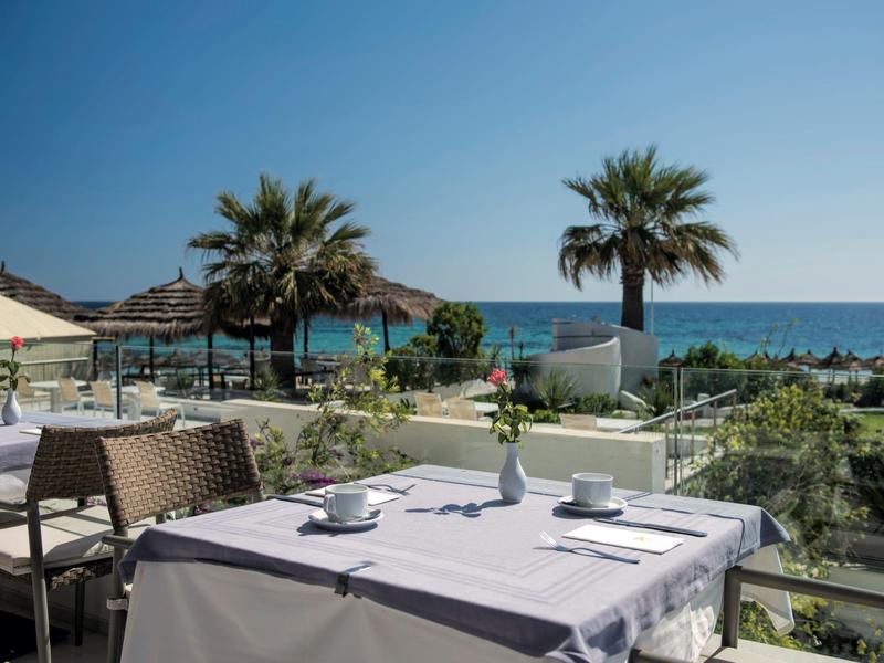 Terrazza con tavolo apparecchiato con vista su palme e mare sotto un cielo sereno.