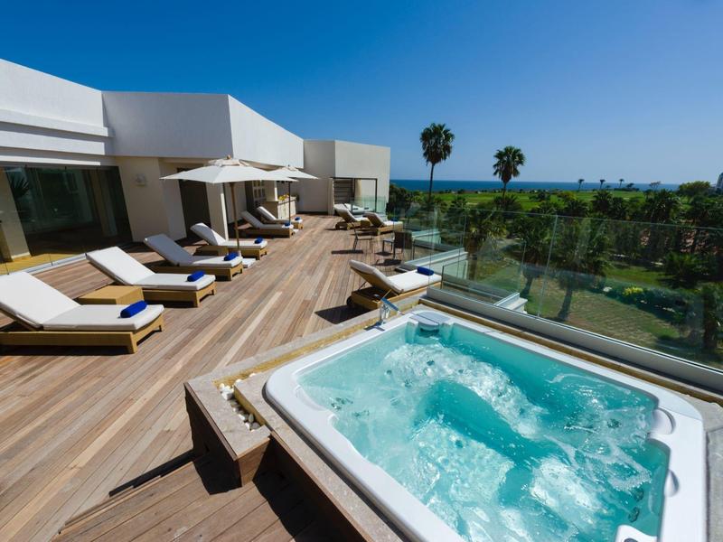 Terrazza con vasca idromassaggio, lettini prendisole e vista su un giardino con palme e mare sullo sfondo.