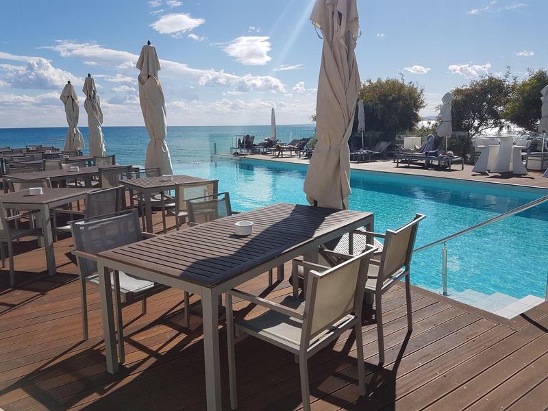 Espace piscine avec tables, chaises et vue sur la mer par temps ensoleillé.