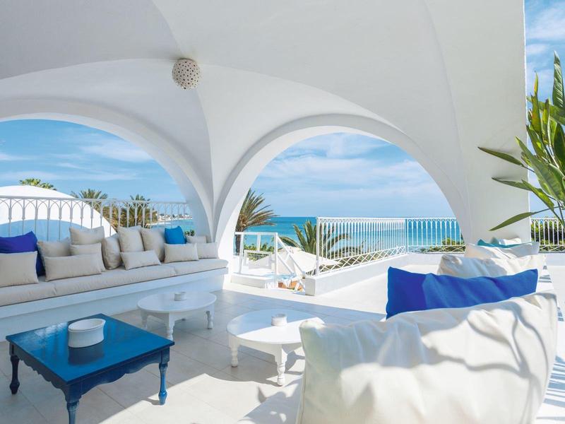Terrasse en bord de mer lumineuse avec arches blanches, table bleue, sièges confortables et plantes luxuriantes.