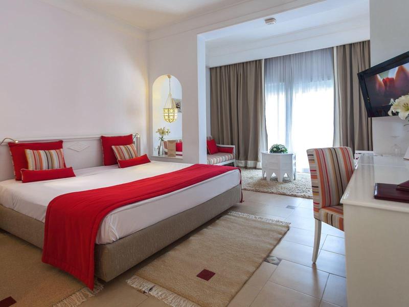 Chambre d'hôtel moderne avec grand lit, accents rouges et balcon.