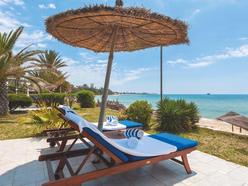 Deux chaises longues aux coussins bleus sous un grand parasol en paille près de la plage.