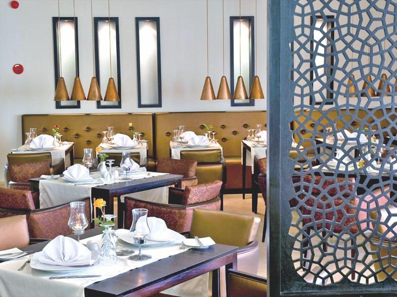 Modern eingerichteter Restaurantbereich mit hellen Stühlen, weißen Tischtüchern und dekorativen Spiegelfenstern.