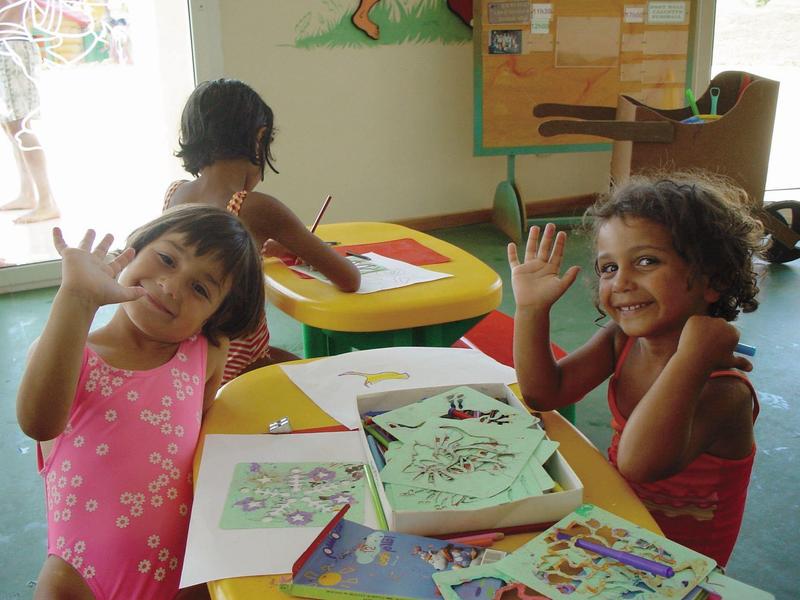 Drei Kinder sitzen an bunten Tischen und winken fröhlich in einem hellen Raum.