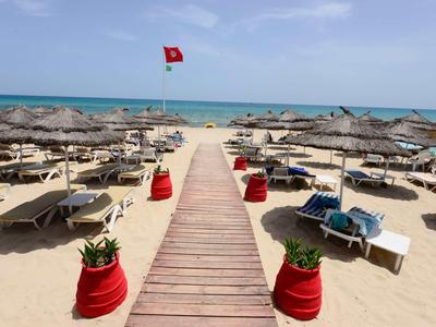 Holzsteg führt durch Sandstrand mit Liegestühlen und Sonnenschirmen zum Meer mit roter Flagge.