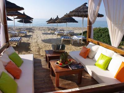 Strandblick mit weißen Sofas, bunten Kissen, Holztisch und parasollen im Sand am Meer.