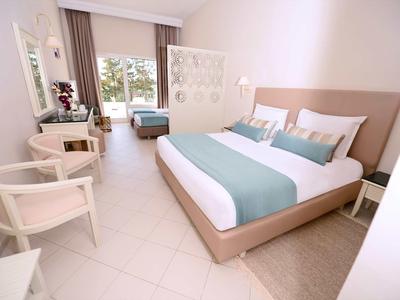 Světý hotelový pokoj s velkou postelí, dvěma židlemi, psacím stolem a balkonem s rostlinami.