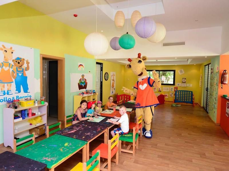 Děti si hrají u stolů v barevné, přívětivé herně s maskotem.