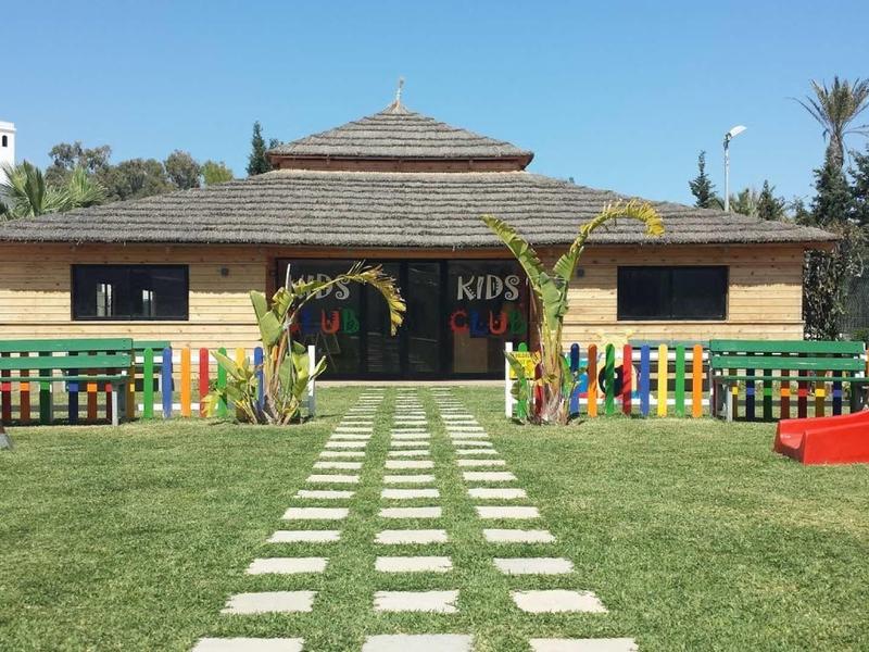 Dětské hřiště s klouzačkou před dřevěnou budovou s barevným plotem a zahradní cestičkou.