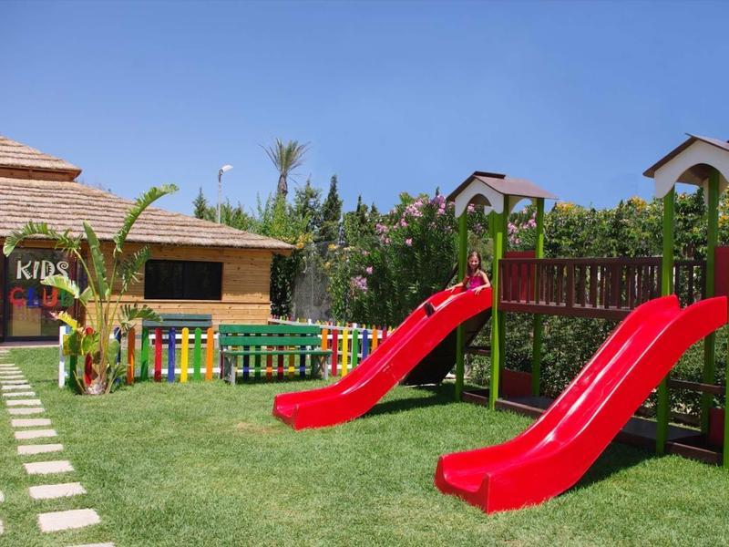 Dětské hřiště s červenými skluzavkami a barevným plotem na zeleném trávníku pod modrou oblohou.