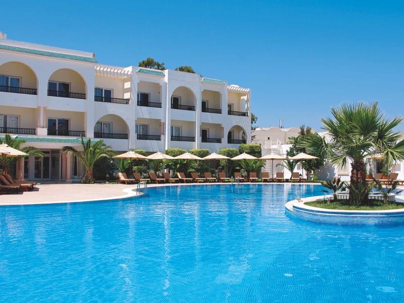 Velký hotelový bazén s deštníky a palmami před bílou budovou pod jasně modrou oblohou.