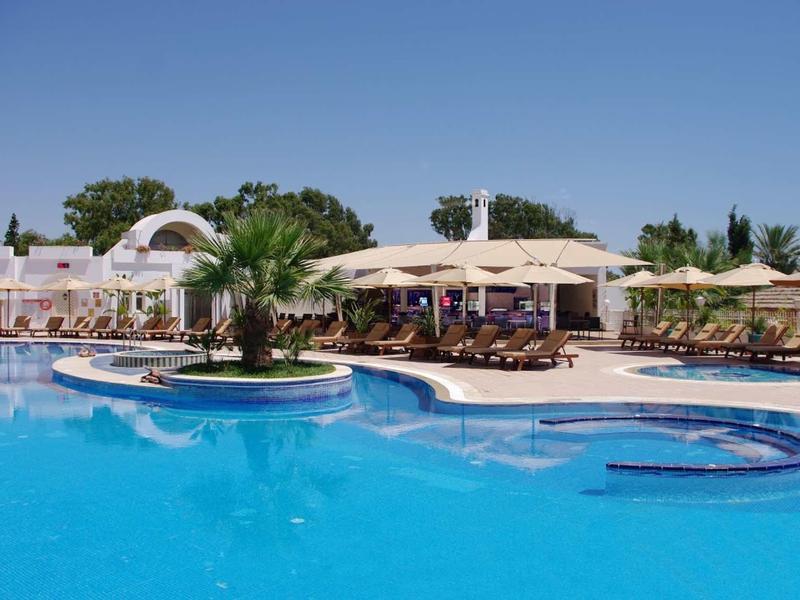 Velký venkovní bazén s palmami a slunečníky vedle hotelové budovy pod jasnou oblohou.