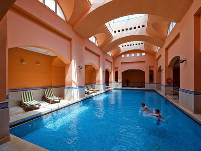 Innenpool mit blauem Wasser, orangefarbenen Wänden und mehreren Liegestühlen am Rand.