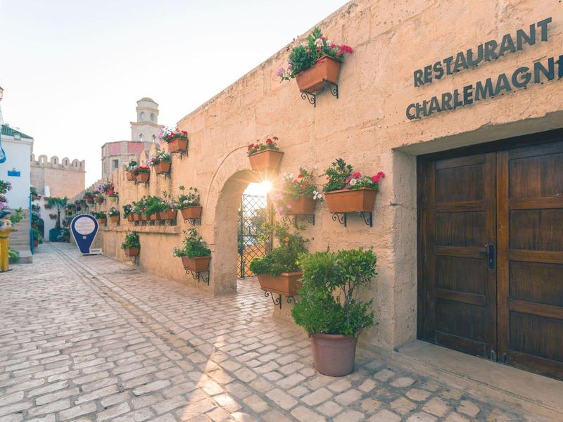 Stretta strada affascinante con vasi di fiori e ingresso del Ristorante Charlemagne alla luce calda del sole.