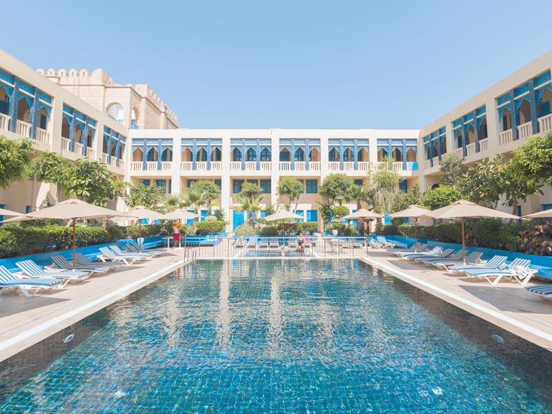 Piscina dell'hotel con lettini blu e ombrelloni, circondata da un edificio moderno sotto un cielo sereno.