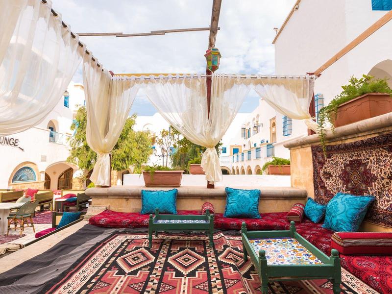 Accogliente lounge esterna con cuscini decorati, tavolini bassi e tende bianche leggere sotto un cielo limpido.