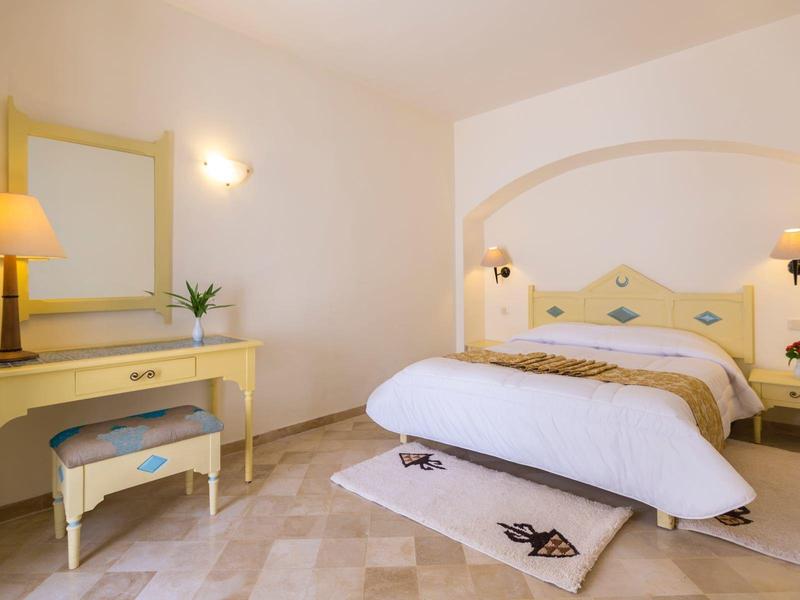 Camera d'hotel luminosa con letto grande, comodini, specchio e lampada in tonalità gialle morbide.