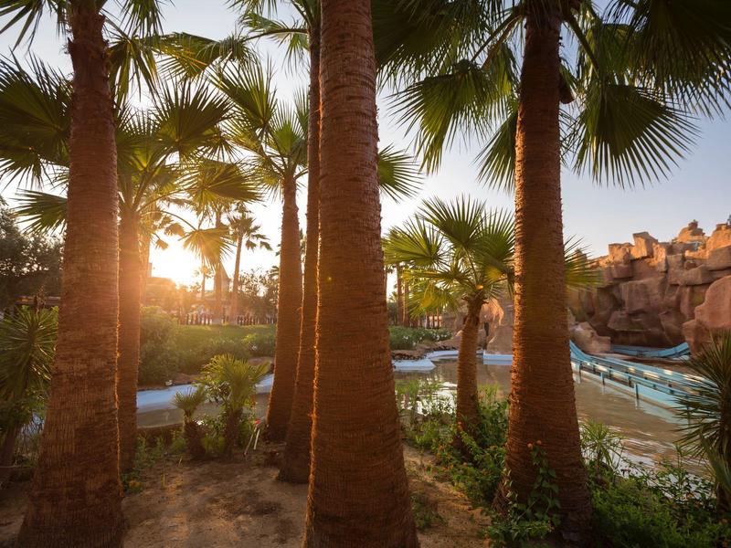 Tramonto visto attraverso palme, con vista su un parco acquatico con scivoli e strutture rocciose.