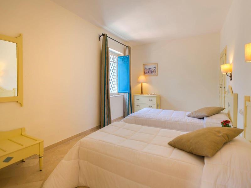 Camera d'hotel luminosa con due letti singoli, mobili gialli e finestra aperta con tenda blu.