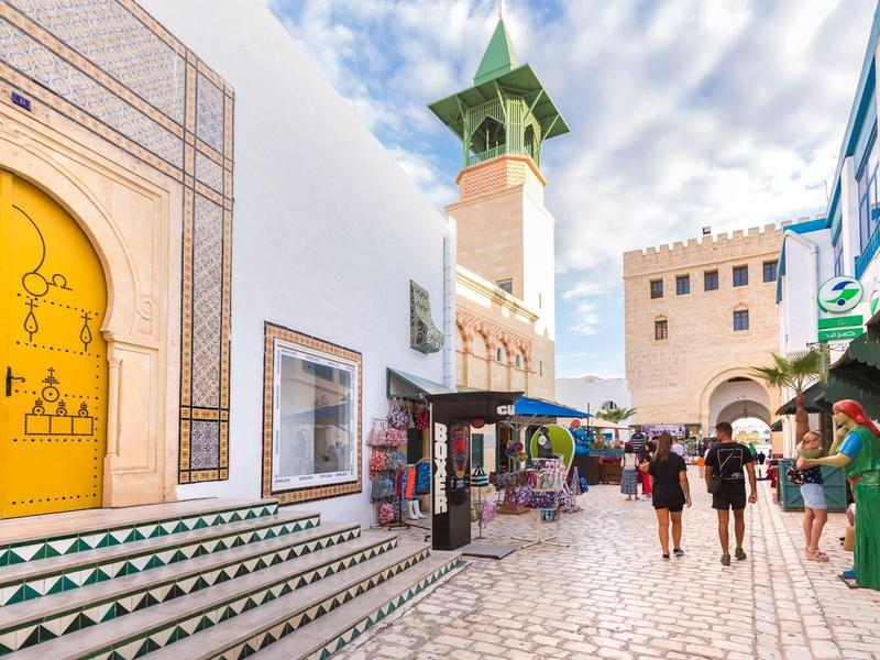 Scena di strada con persone che camminano vicino a edifici bianchi e colorati e una torre alta sotto un cielo parzialmente nuvoloso.