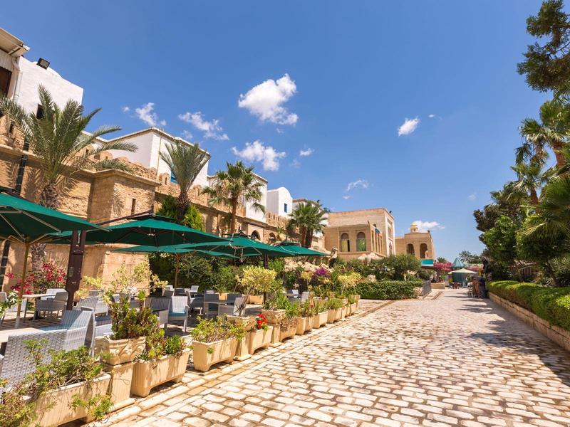 Area pranzo all'aperto con tavoli e ombrelloni lungo un vialetto lastricato vicino a un hotel.