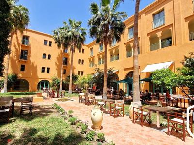 Innenhof mit orangem Gebäude, Palmen, Garten und Holztischen mit Stühlen unter Sonnenschirmen.