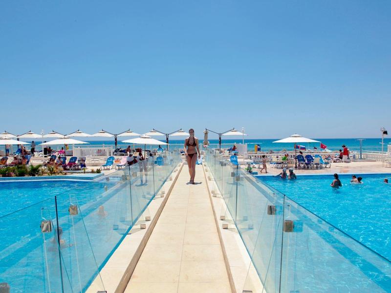 Mujer camina por un muelle entre dos piscinas con vista al mar y cielo azul.