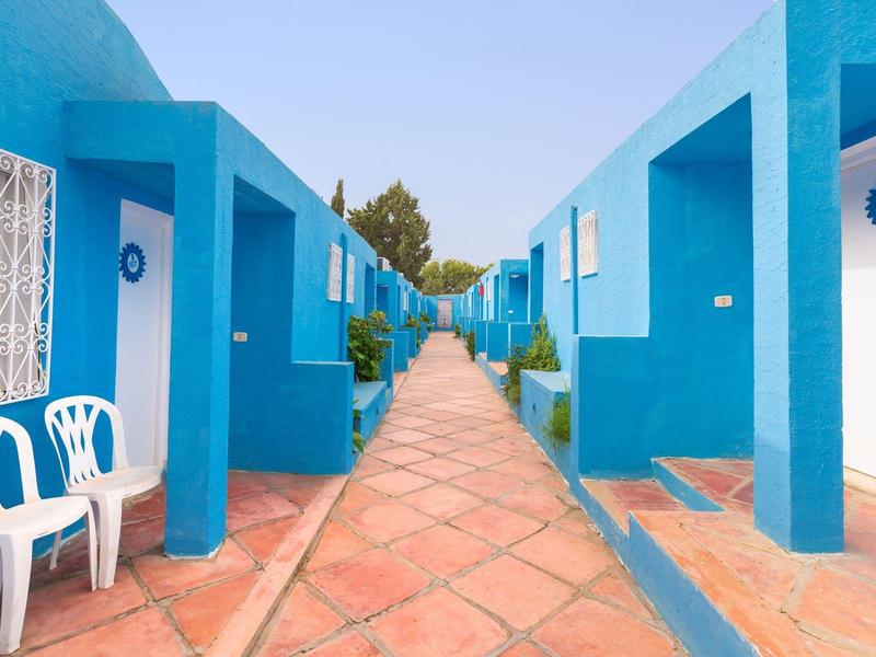 Habitaciones de hotel azules con puertas abiertas y baldosas de terracota en el exterior bajo cielo despejado.