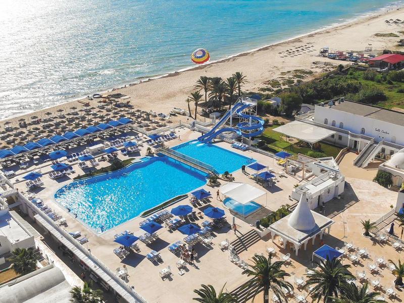 Resort frente a la playa con piscina grande, sombrillas y áreas de descanso junto al mar.
