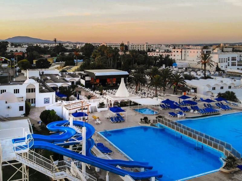 Área de piscina del hotel con toboganes acuáticos, tumbonas azules y edificios alrededor al atardecer.