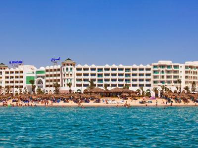 Großes Hotel am Strand mit Sonnenschirmen, Palmen und blauem Meer unter klarem Himmel.