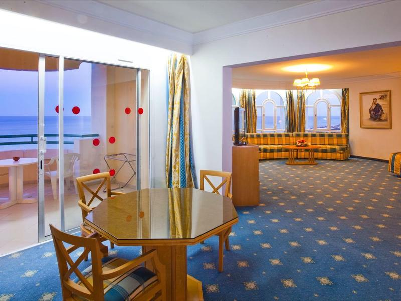 Geräumiges Hotelzimmer mit Sitzbereich, Balkon und Blick auf das Meer bei Sonnenuntergang.