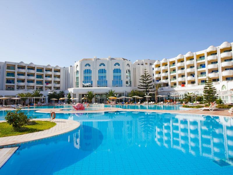 Großes Hotel mit zwei Flügeln und einem Pool im Vordergrund unter klarem blauem Himmel.