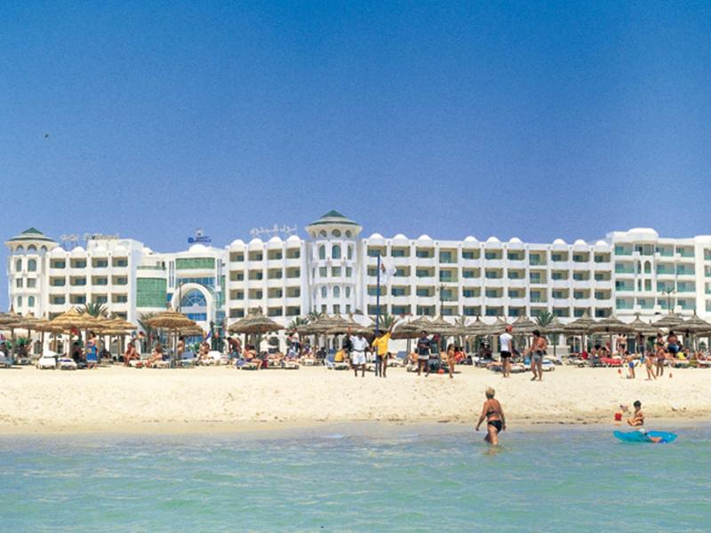 Großes Hotel am Strand mit Menschen im Wasser und Sand unter blauem Himmel.