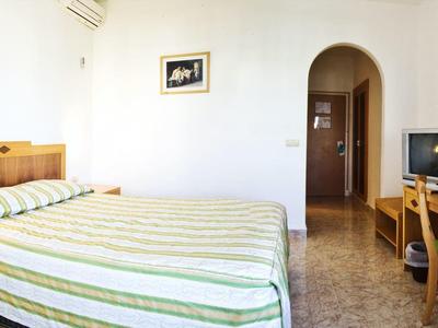 Světle dvoulůžkový pokoj s pruhovanou postelí, nočním stolkem, psacím stolem a velkým zrcadlem.