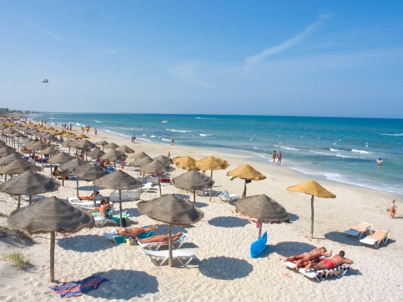 Pláž s mnoha slunečníky a lehátky, v pozadí moře pod jasnou oblohou.