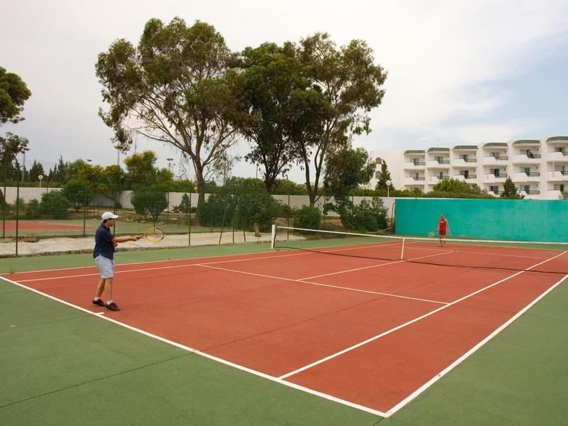 Dva lidé hrají tenis na venkovním kurtu vedle bílé hotelové budovy.