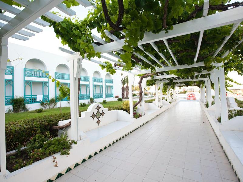 Krytá cesta s bílými pergolami a zelenými révami vedle hotelové budovy.