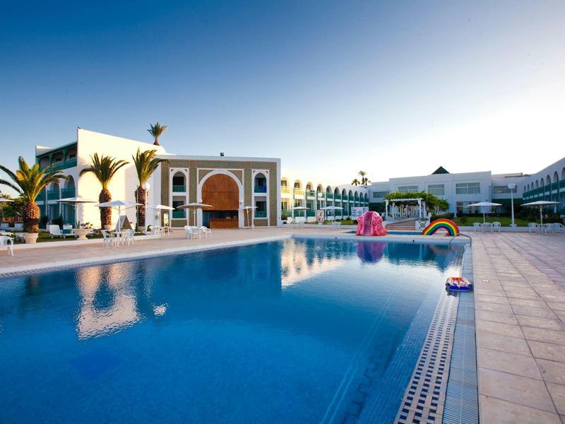 Velký hotelový bazén s čistou vodou obklopený budovami lemovanými palmami pod modrou oblohou.