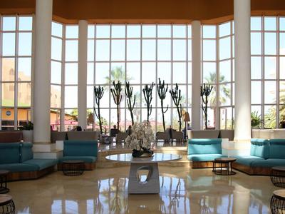 Große Hotellobby mit hohen Fenstern, Sitzbereichen und sonnigem Blick nach draußen.