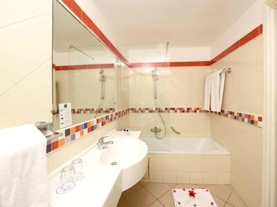 Světlá koupelna s vanou, velkým zrcadlem a bílou keramickou výbavou, barevné dlaždicové akcenty.