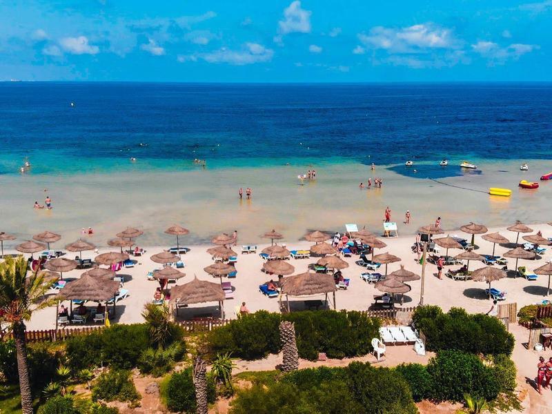 Pláž s deštníky, lehátky a hosty u modrého moře pod jasnou oblohou.