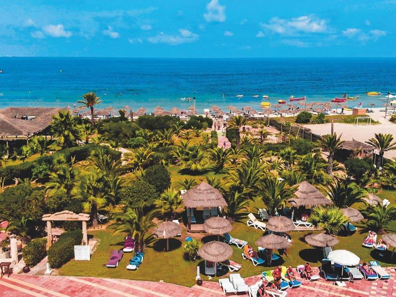 Pohled na letovisko se zelenými palmami, slunečními parasoly a modrým mořem v pozadí.