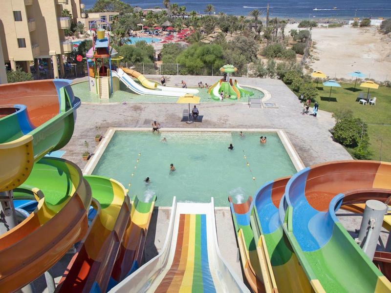 Barevné vodní skluzavky vedoucí do hotelového bazénu s výhledem na pláž.