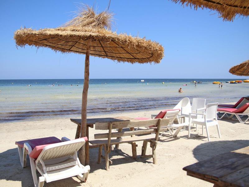 Písčitá pláž se slámovými slunečníky, lehátky a klidným mořem v pozadí.